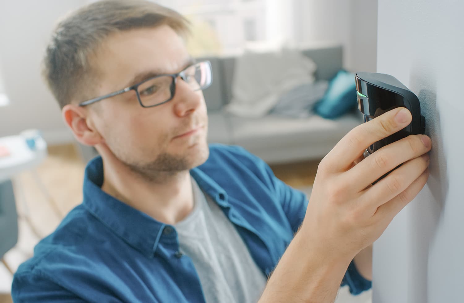 Young Man in Glasses Wearing a Blue Shirt is Hanging a Modern Movement Detector Unit on a White Wall at Home